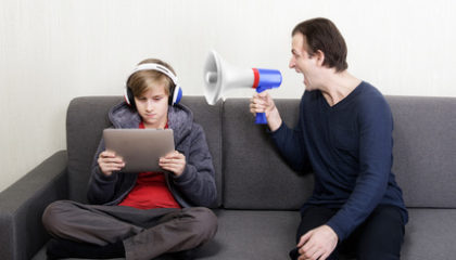 Tween son in headphones looks at the digital tablet display while his father yells at him through a megaphone